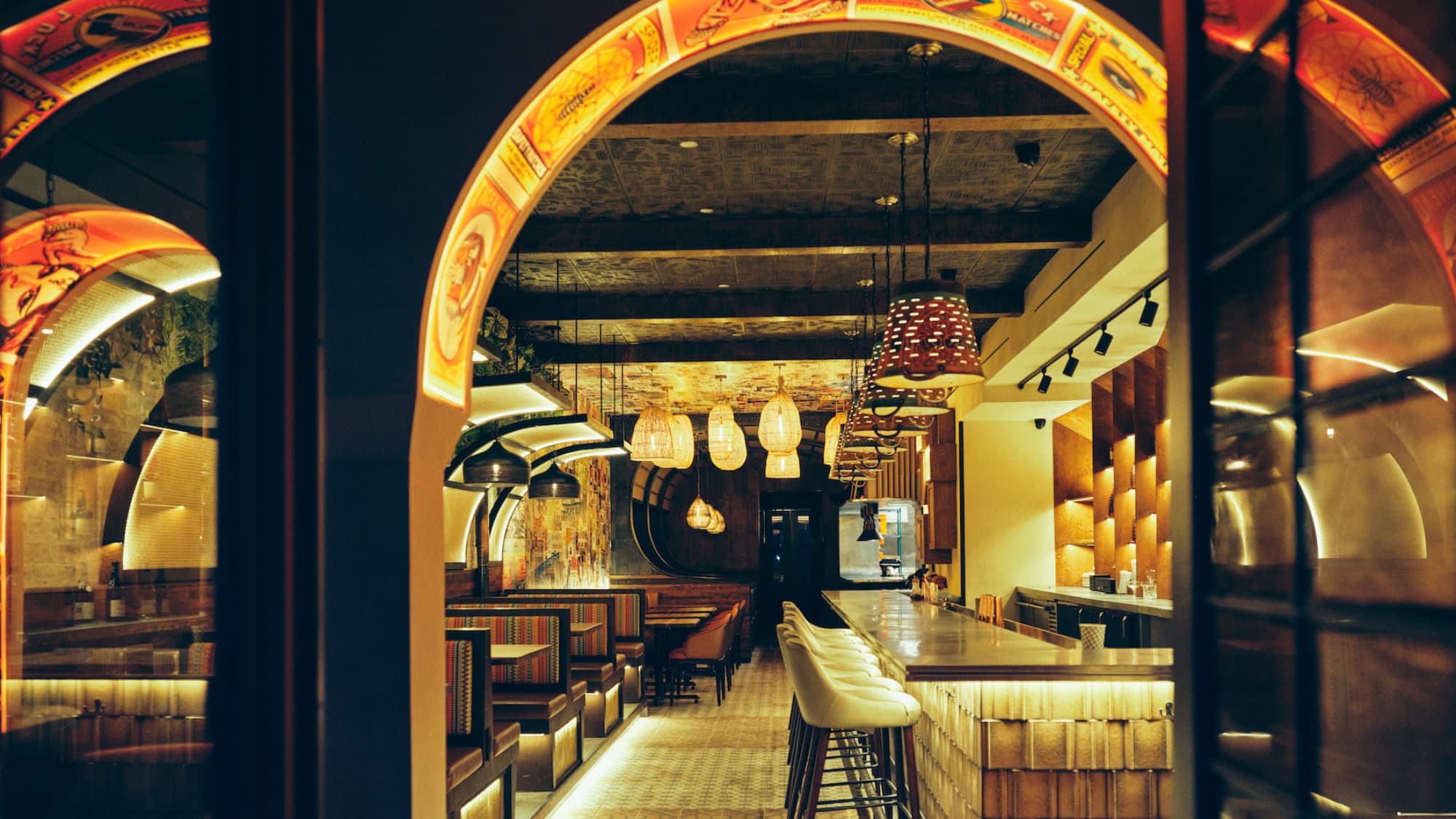 East Village Adda dining room through a matchbook-lined arch: striped booths, rattan pendants, and a glowing bar