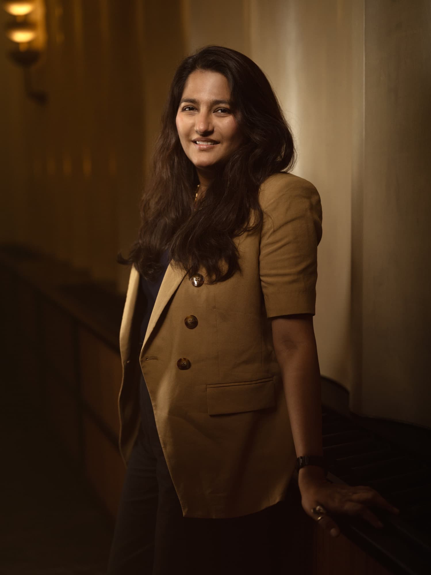 Aditi Dugar, founder of Masque, in a tan blazer leaning against a wooden ledge in a dim restaurant interior