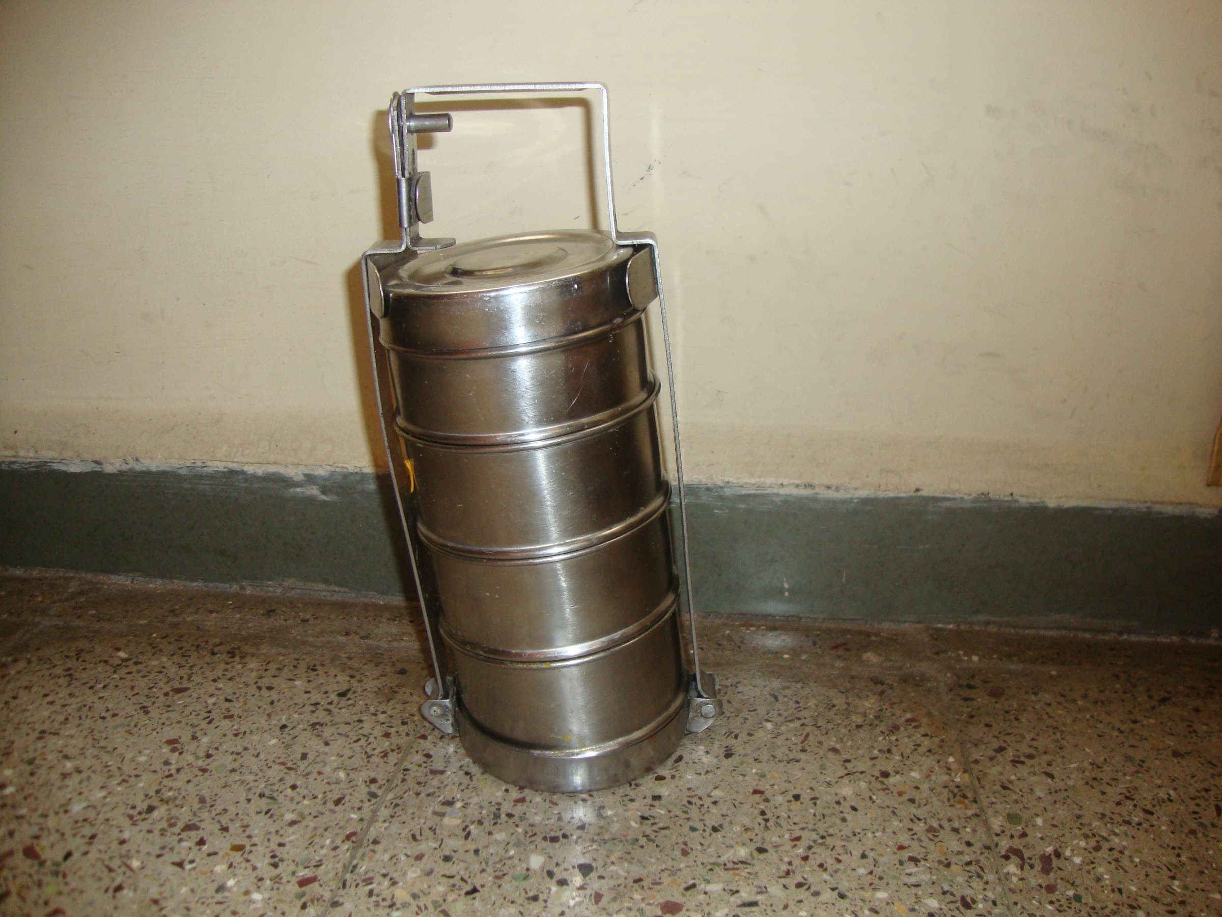 Four-tier stainless steel tiffin carrier (dabba) standing on a terrazzo floor