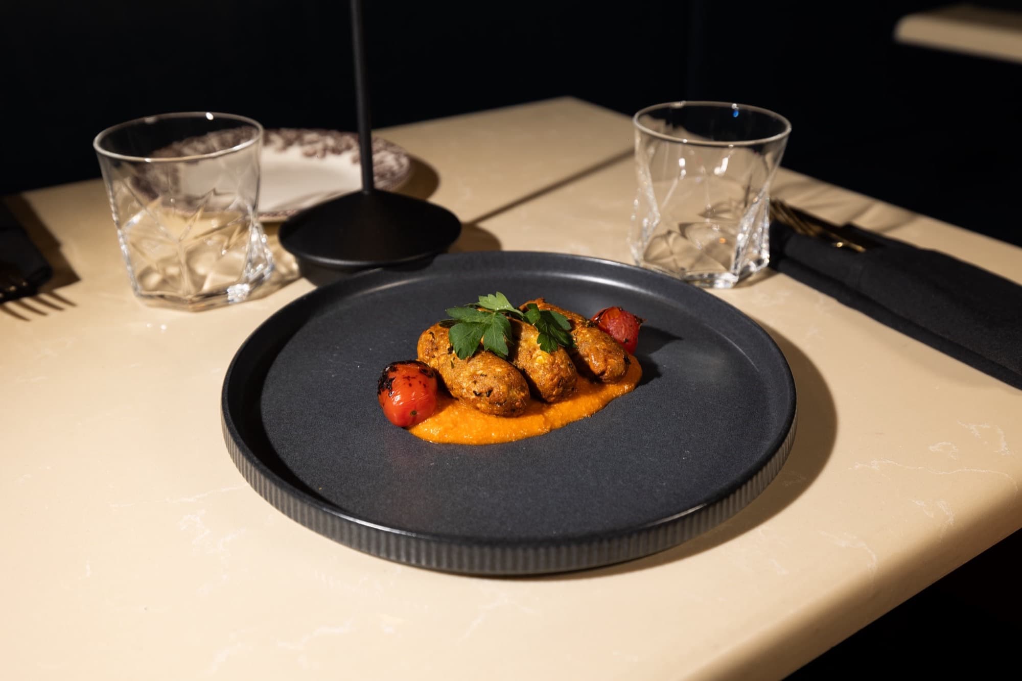 Three golden tikki pieces on orange sauce with blistered tomatoes on a black plate at Badshah Social