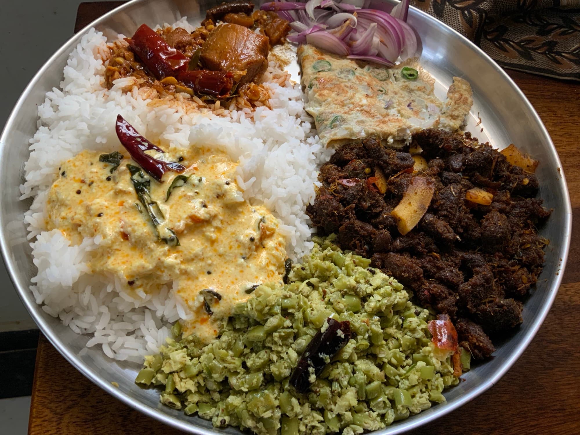 Kerala-style steel thali with rice, coconut vegetable thoran, yogurt curry, dark meat fry, omelette, pickle, and onion