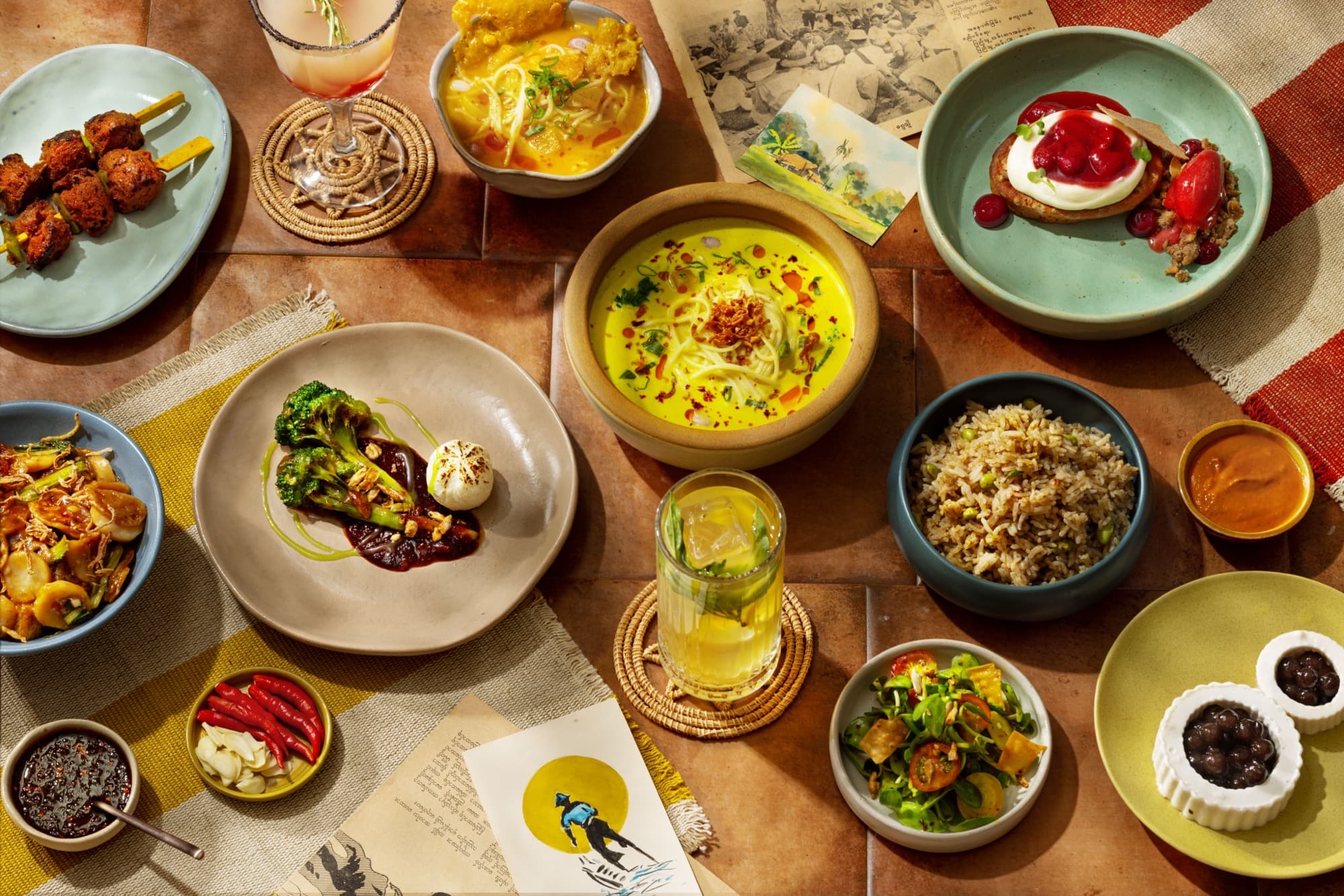 Overhead spread of Burma Burma plates: coconut khao suey, tea-leaf salad, fried rice, zero-proof drinks, and Burmese ephemera on tiled wood