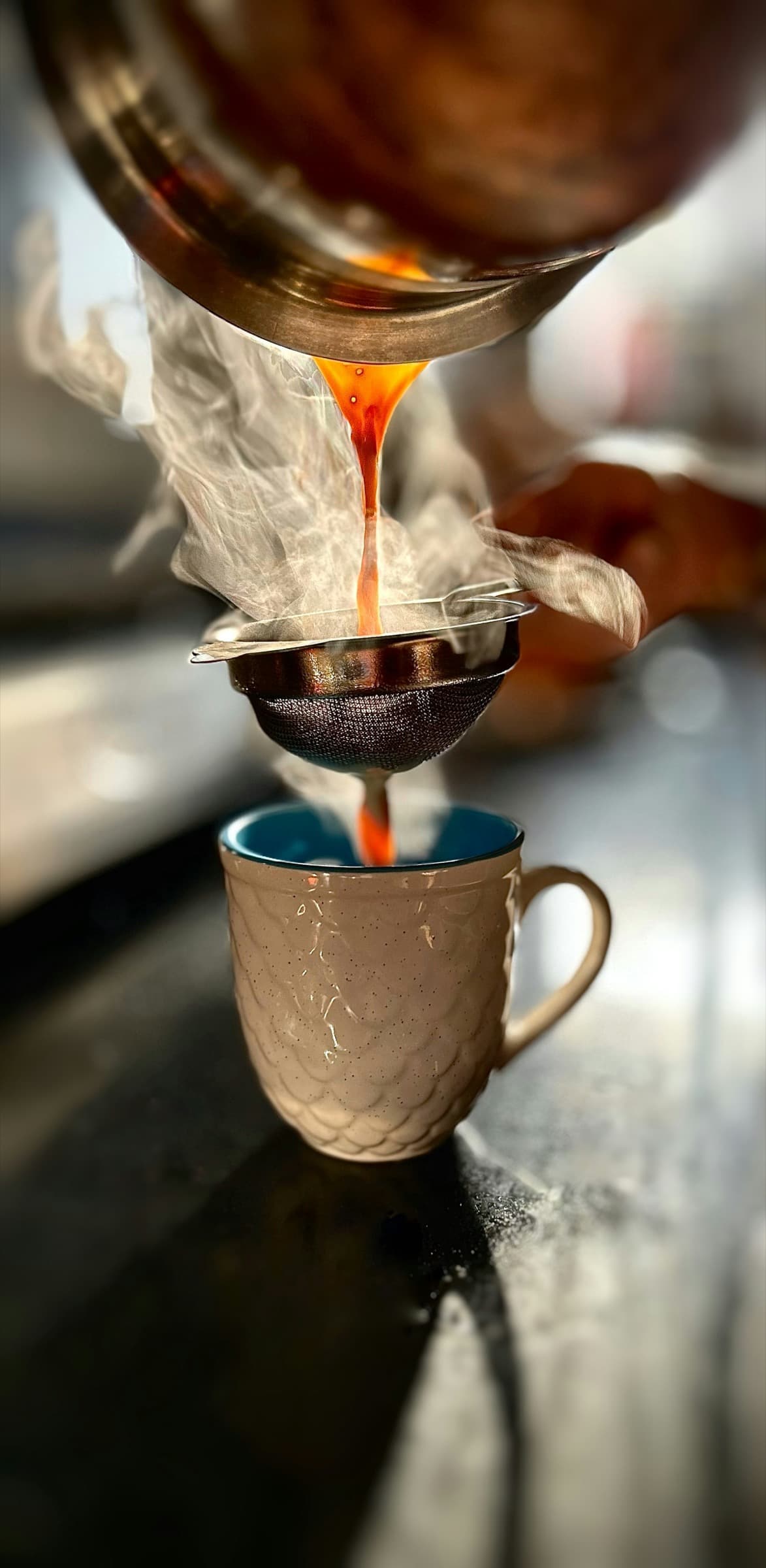 Hot chai poured through a fine mesh strainer into a textured ceramic mug with steam rising