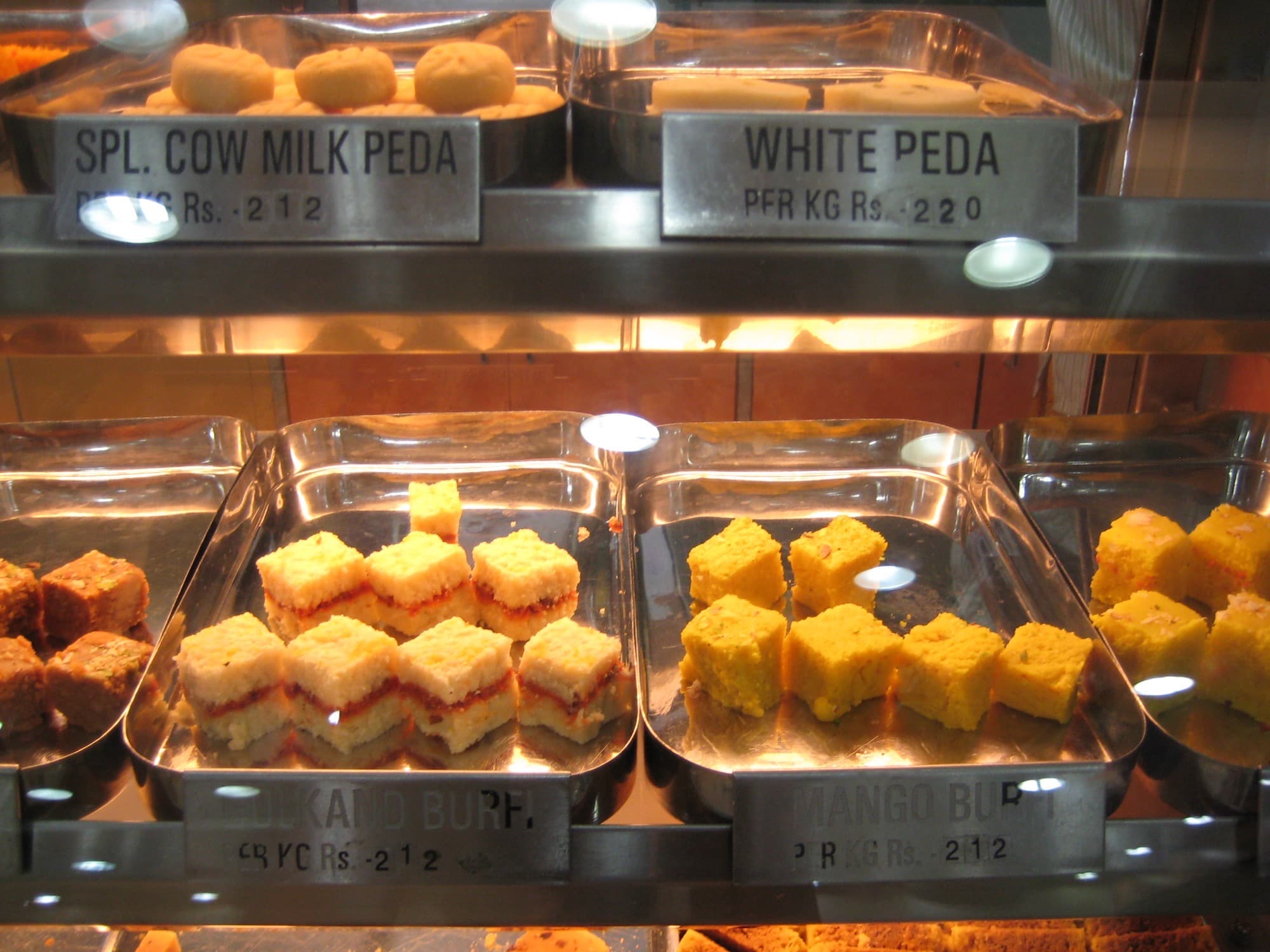 Mithai shop case with trays of cow-milk peda, white peda, gulkand burfi, and mango burfi