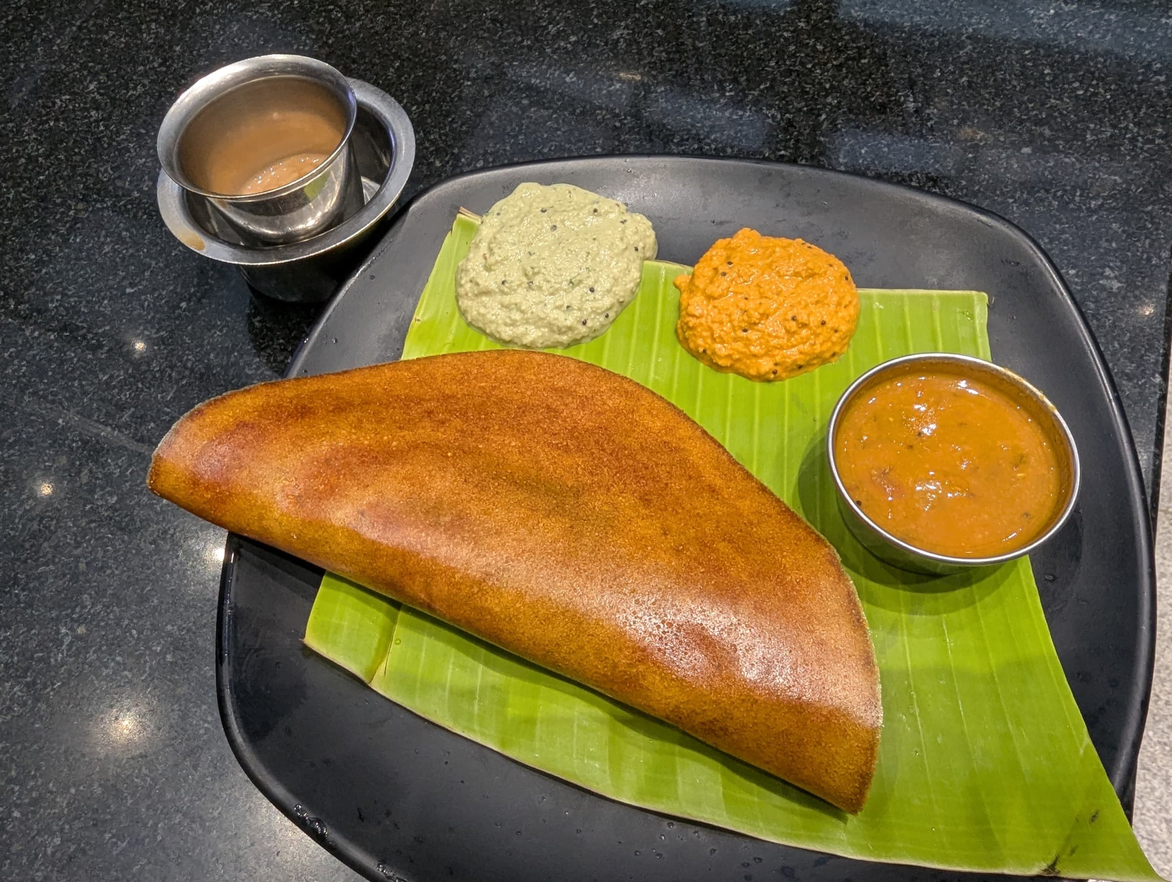 Crispy dosa on banana leaf with coconut and tomato chutneys, sambar, and filter coffee in a steel tumbler