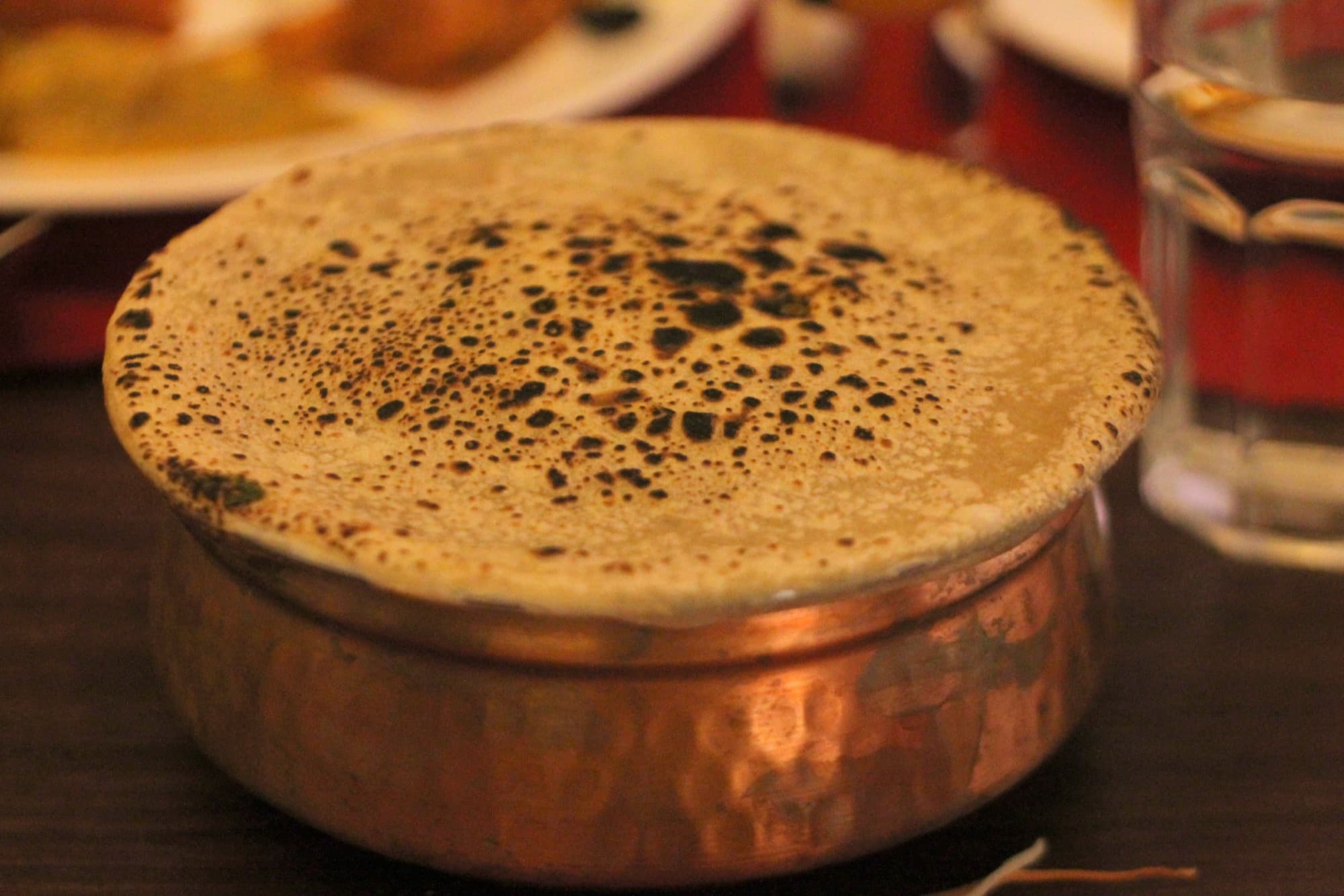 A copper handi covered by a round of flour dough, the surface speckled with dark toasted spots