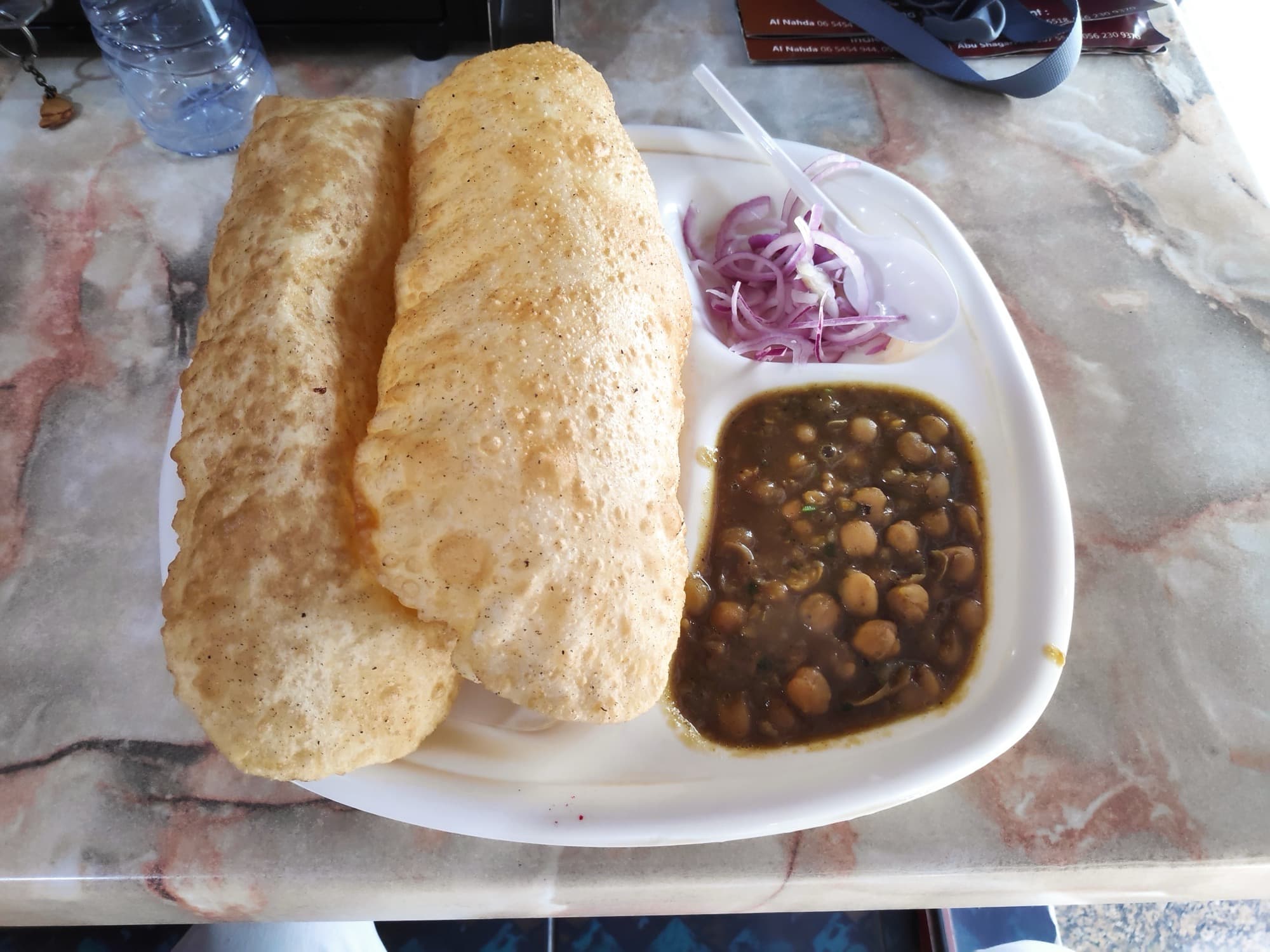 Two puffed bhature beside a bowl of dark chole and sliced red onion on a compartment plate