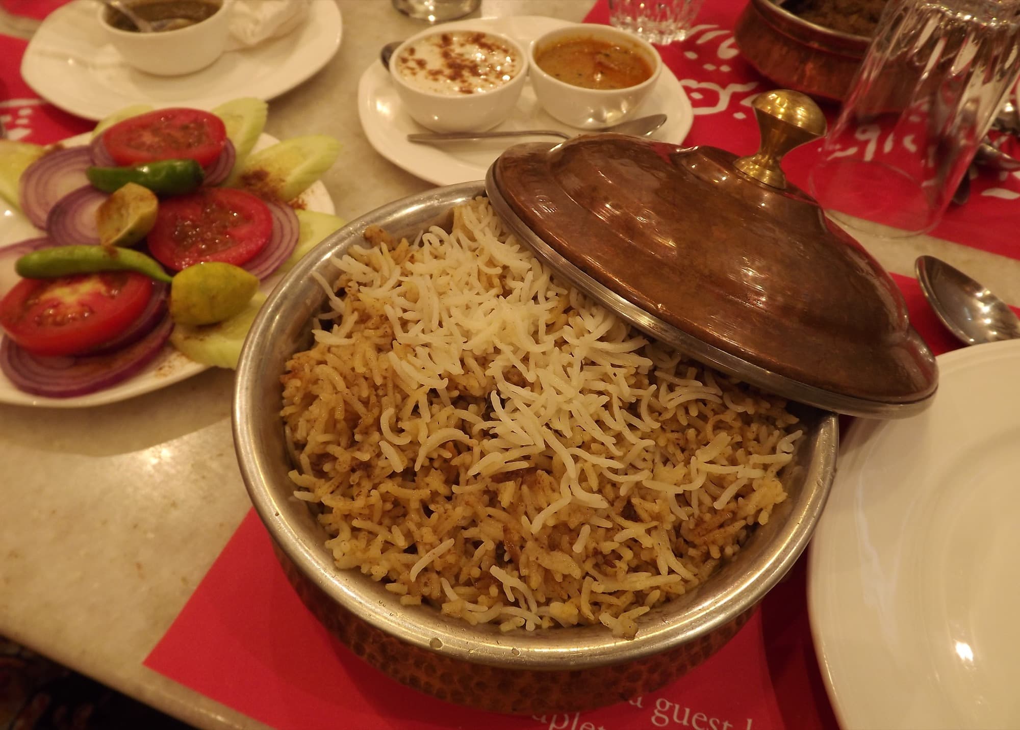 Hyderabadi biryani in a copper handi with the lid off, beside raita, mirchi ka salan, and a tomato-onion salad