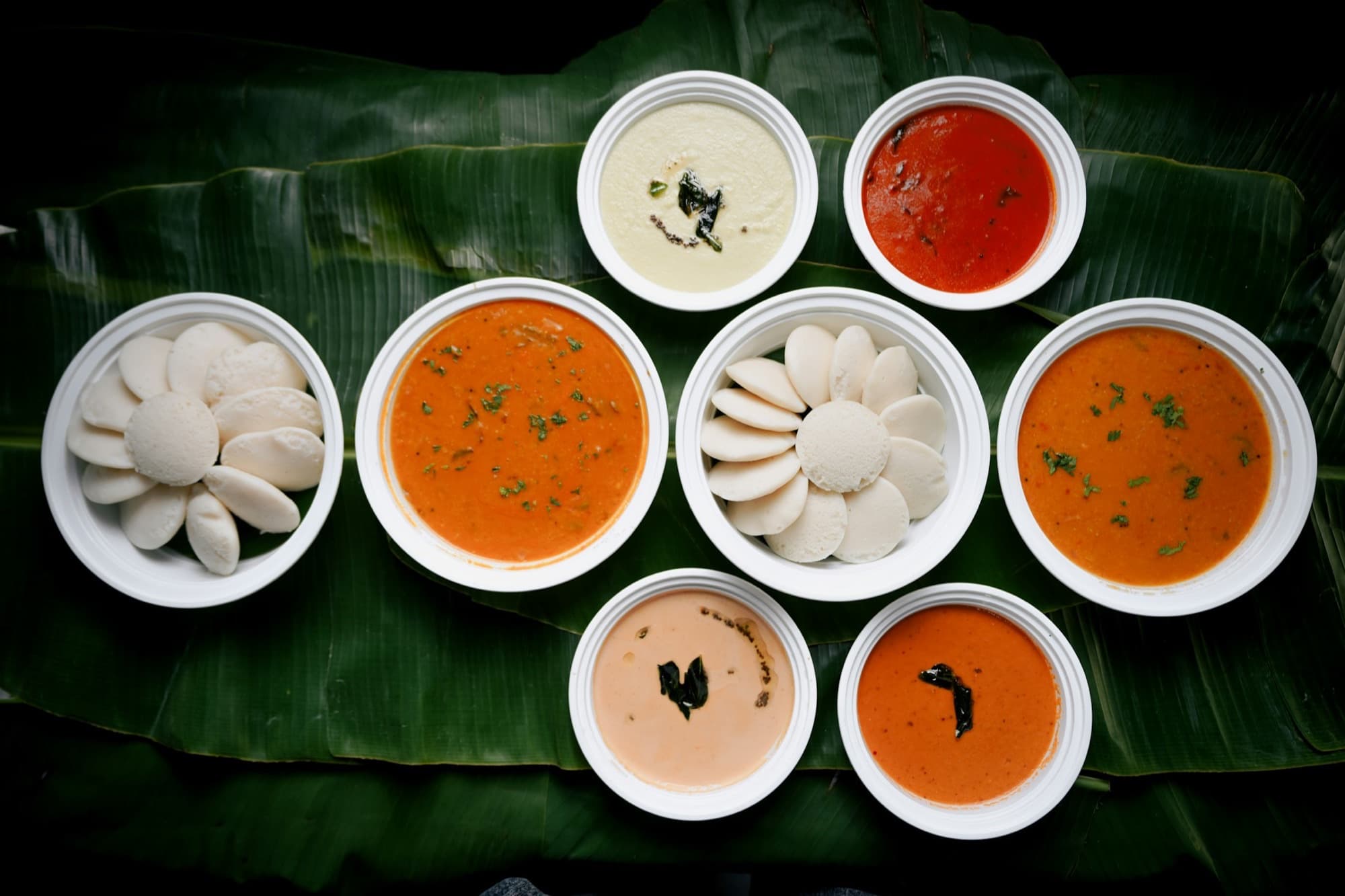 Idli, sambar, and four chutneys in bowls arranged on a banana leaf
