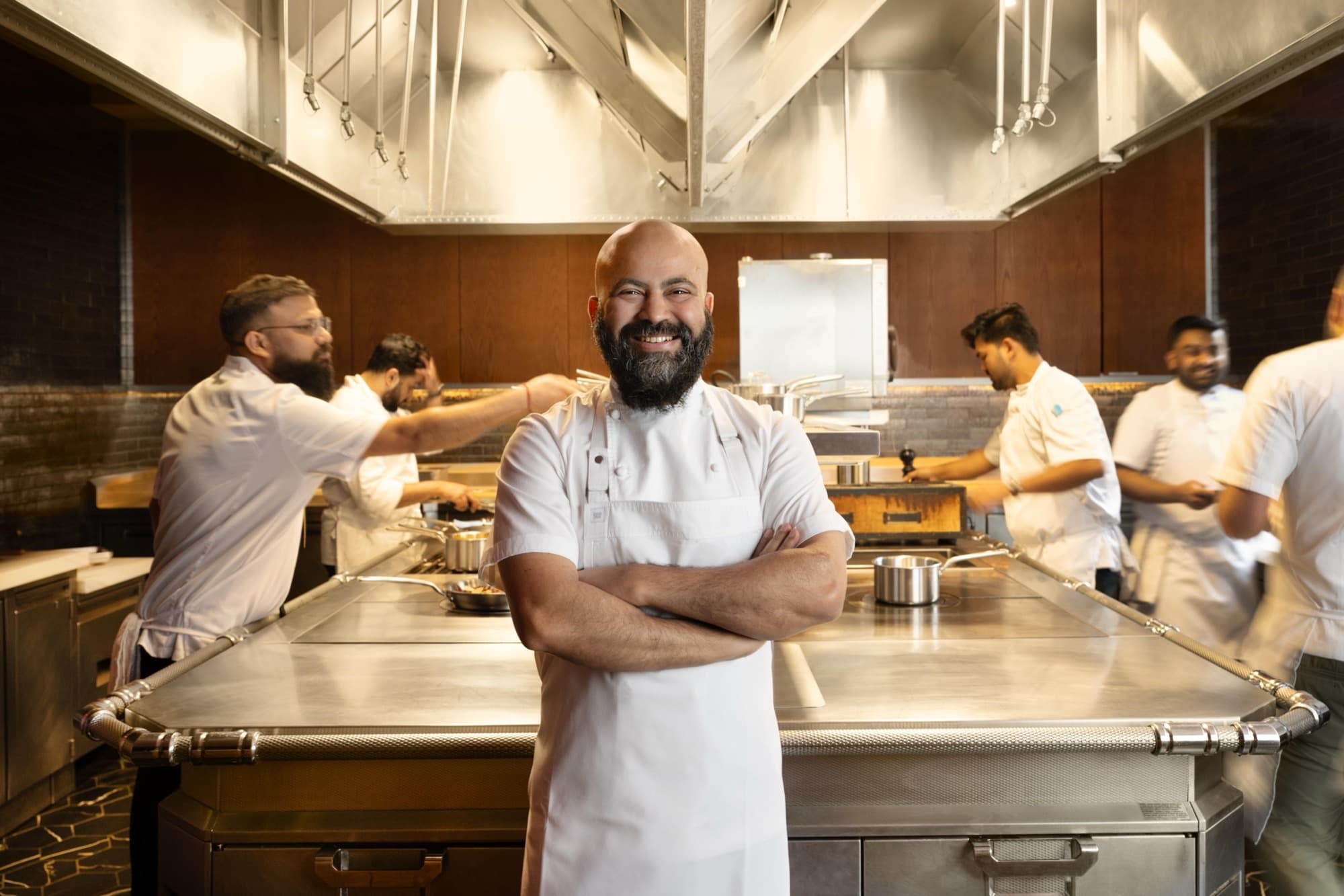 Chef Sujan Sarkar smiling with arms crossed in a white chef’s coat in an open kitchen