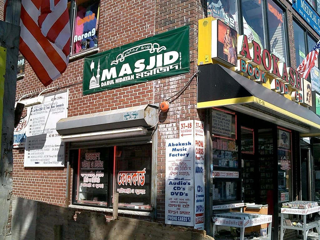 Bangla and English storefronts on 73rd Street in Jackson Heights, Queens, including a bookshop and masjid sign