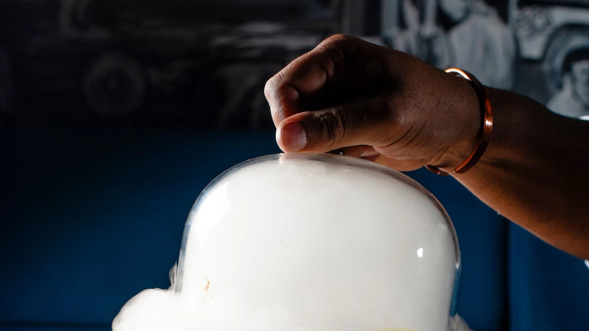 A hand lifts a glass cloche filled with white smoke over a plated dish at Khan Saab