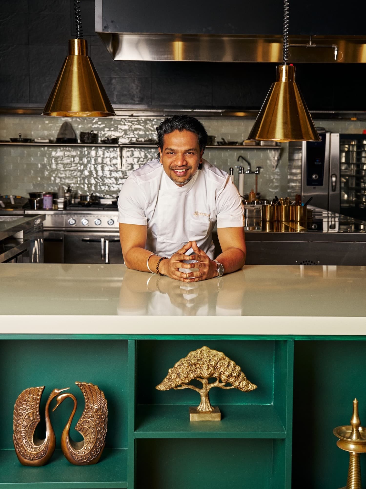 Chef Vinu Raveendran in a white Kidilum coat leaning on the kitchen pass with brass heat lamps behind him