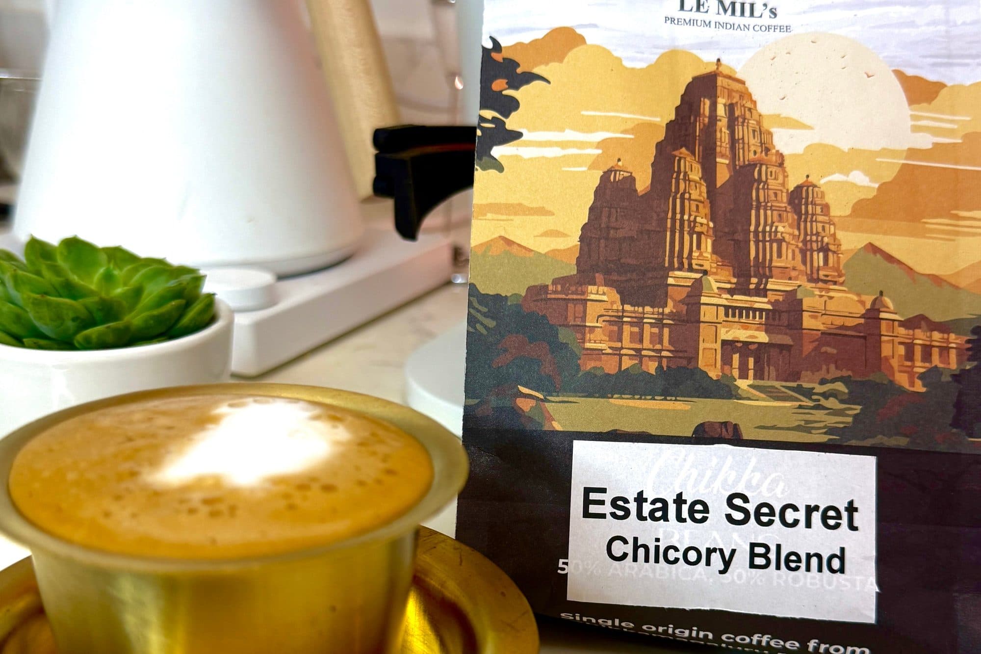 Le Mil’s Estate Secret chicory blend bag beside frothy filter coffee in a brass tumbler