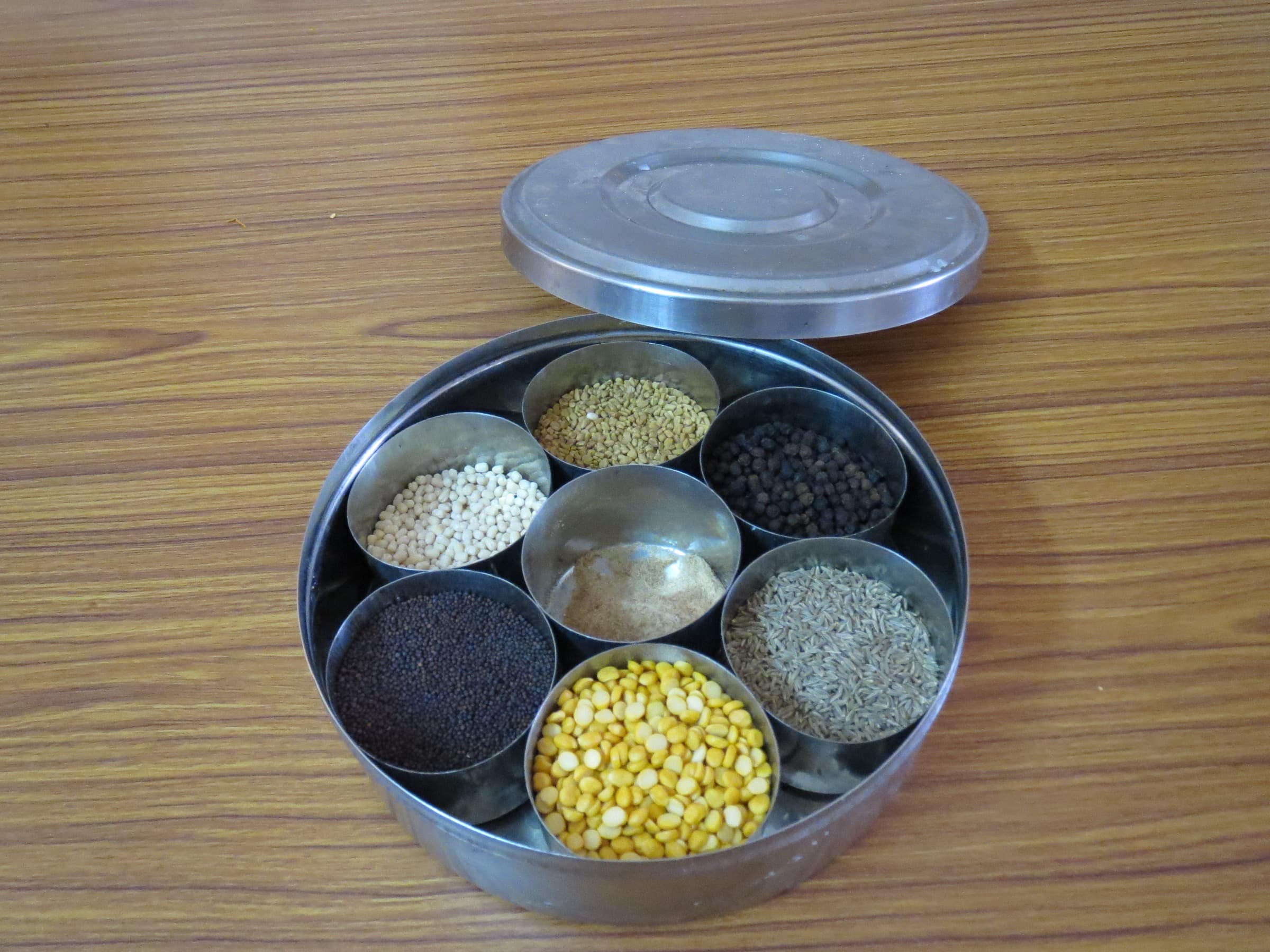 Open stainless anjaraipetti with seven cups of mustard, dals, fenugreek, pepper, cumin, and hing