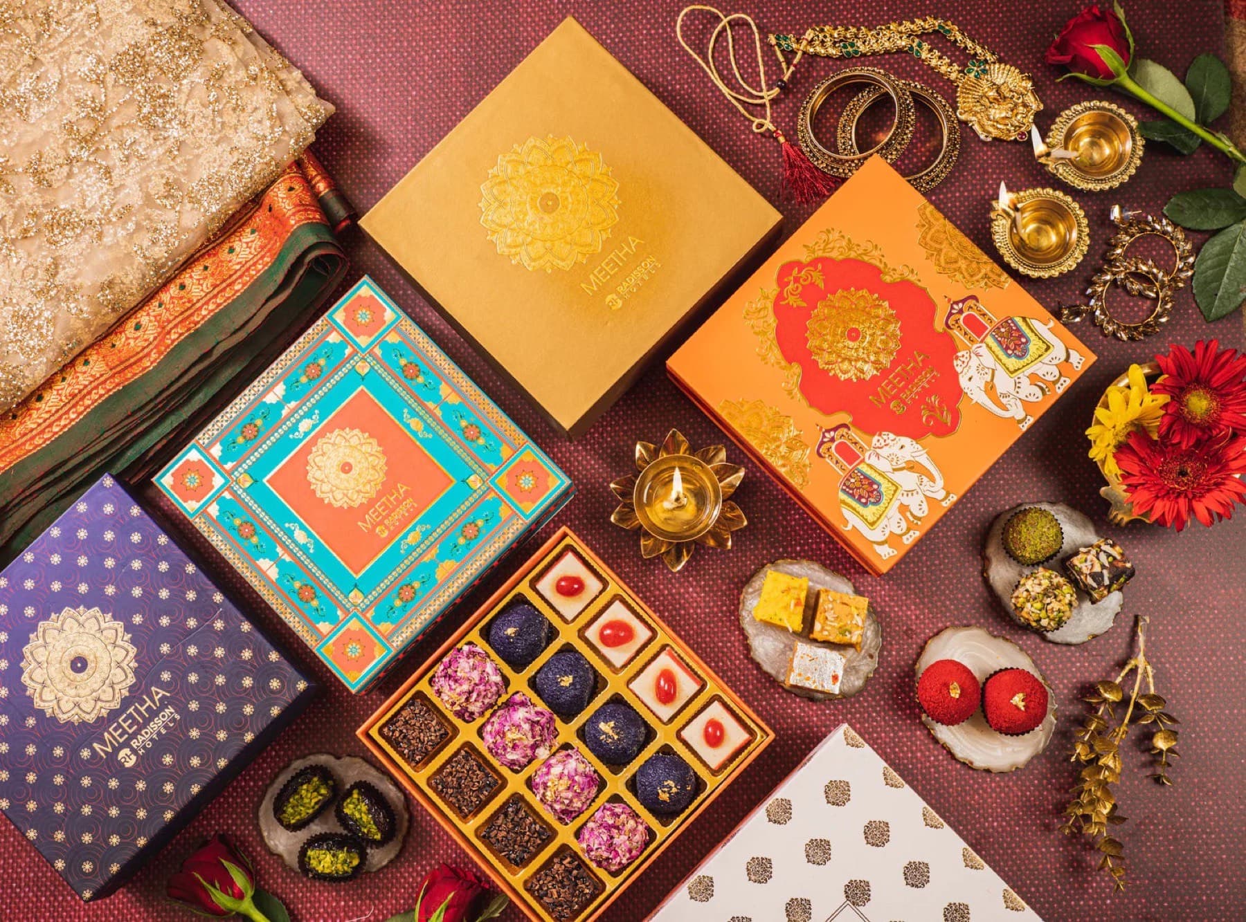 Open Meetha by Radisson mithai gift box surrounded by branded boxes, diyas, roses, and loose sweets