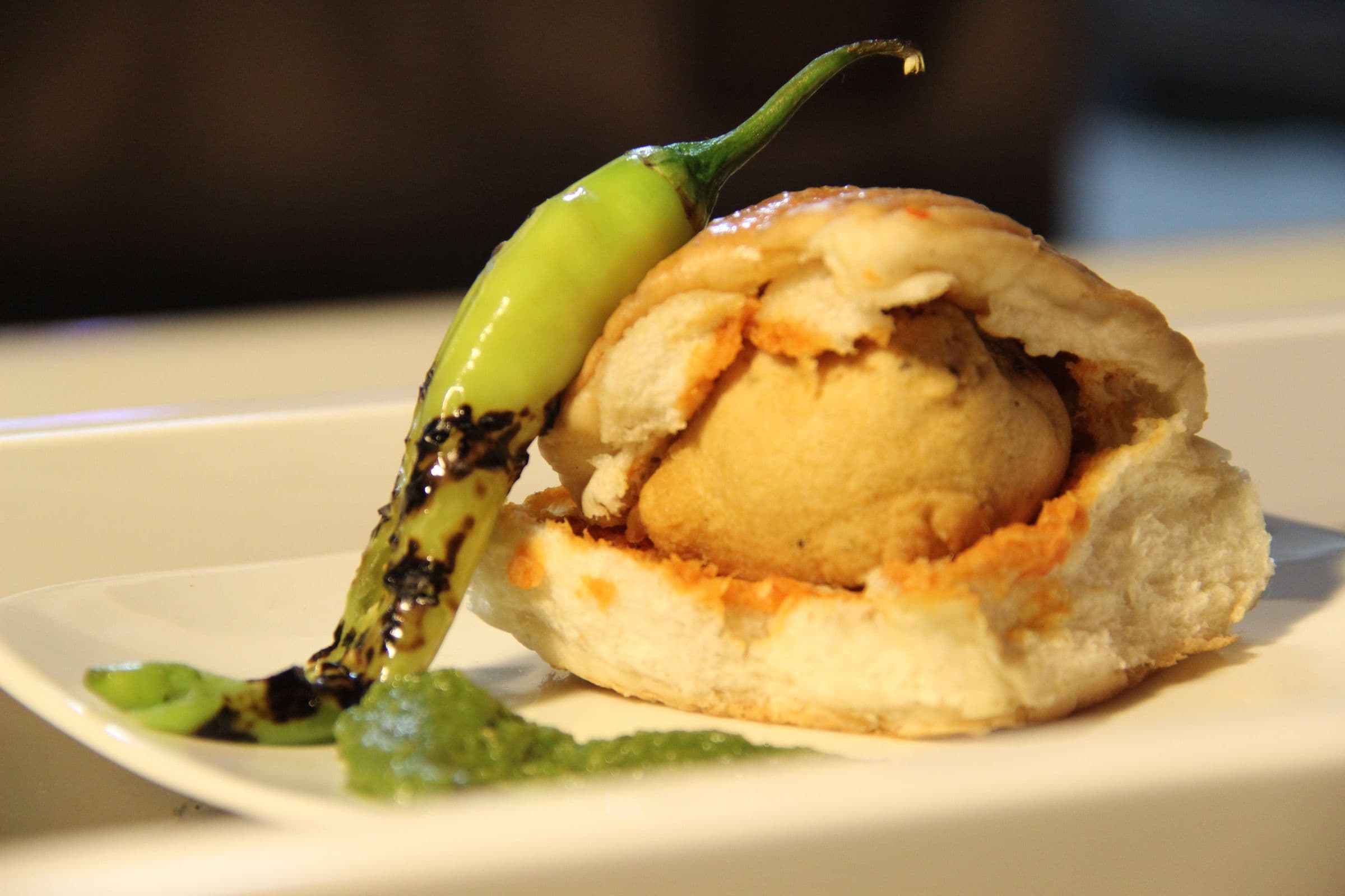 Vada pav on a plate with green chutney and a blistered green chili