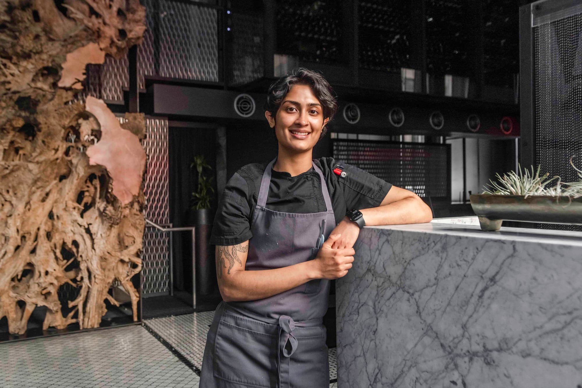 Chef Rasika Venkatesa in a black chef coat and apron, smiling beside a marble counter
