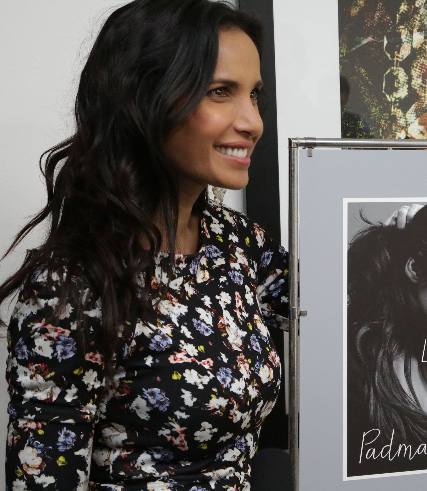 Padma Lakshmi smiling beside a poster for her memoir Love, Loss, and What We Ate