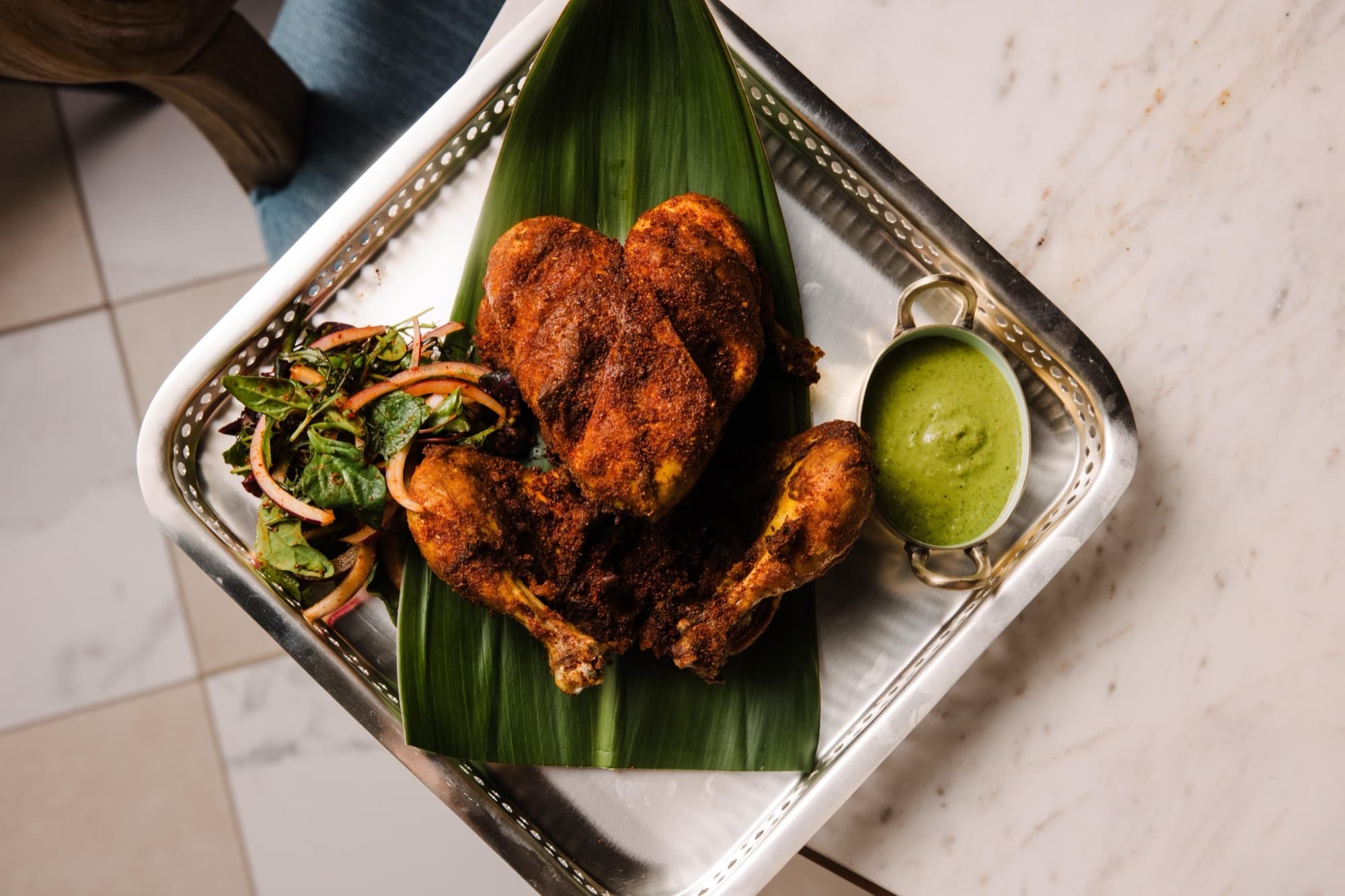 Whole chicken chargha on a banana leaf in a silver tray with green chutney and onion salad