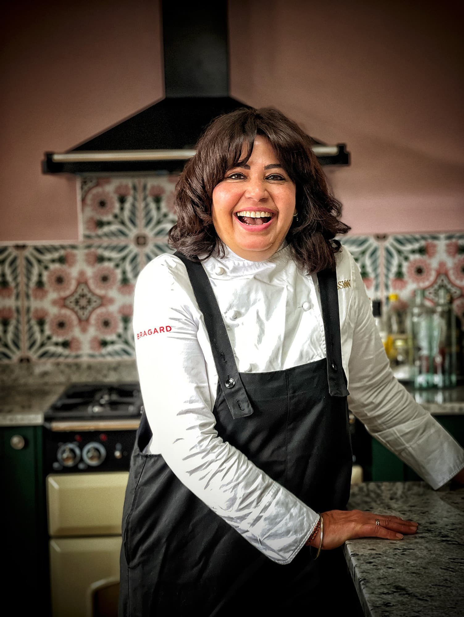 Chef Romy Gill laughing in a white chef’s coat and black apron at a kitchen counter