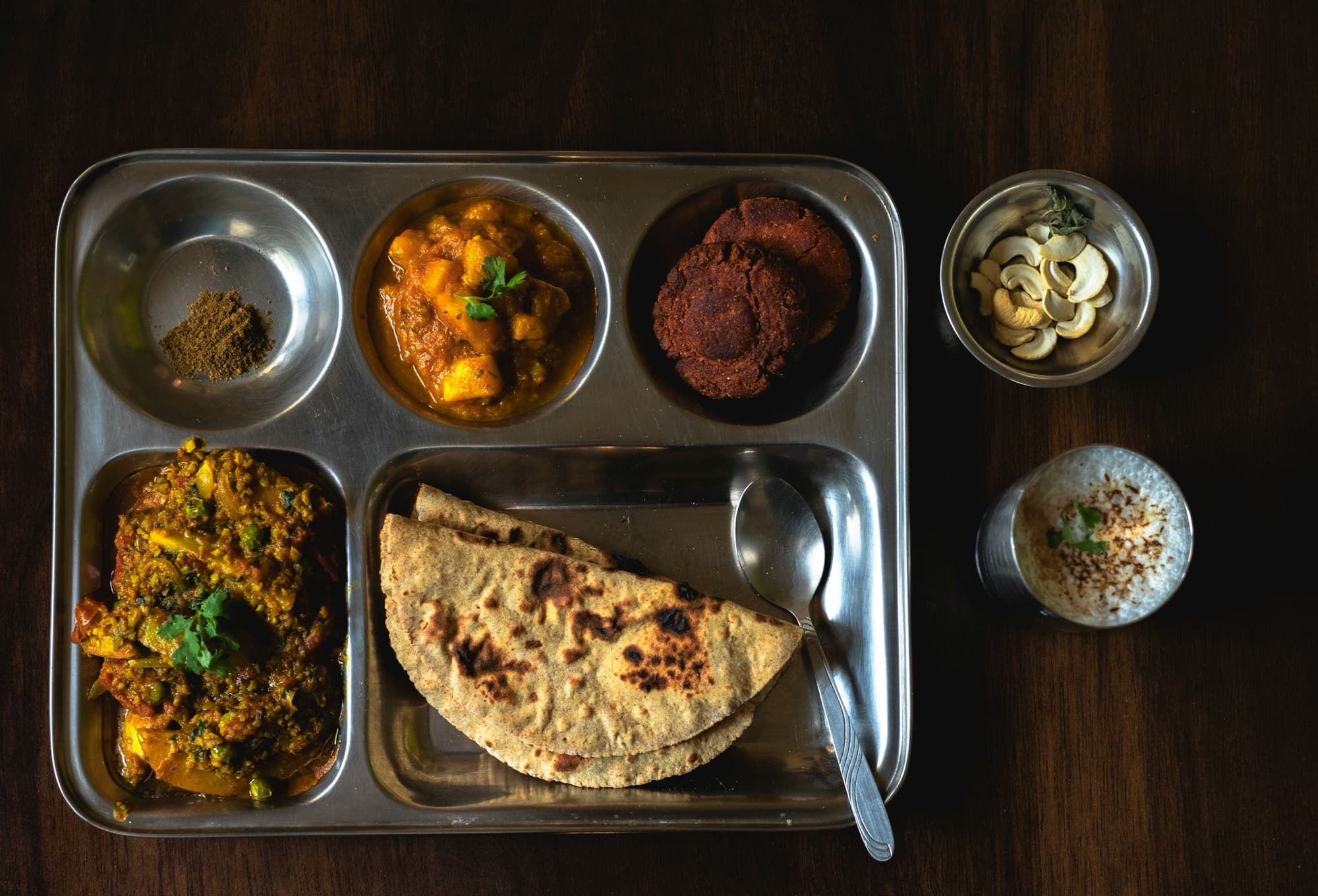 Homemade Indian thali on a metal tray with roti, vegetables, buttermilk, and a sweet