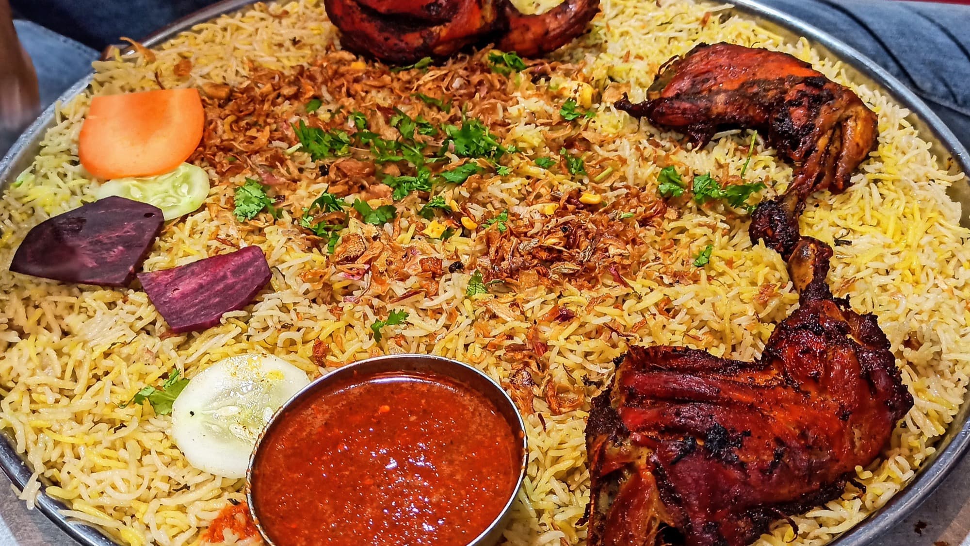 A shared platter of Hyderabadi chicken mandi with roast chicken quarters, fried onions, and a bowl of chile sauce