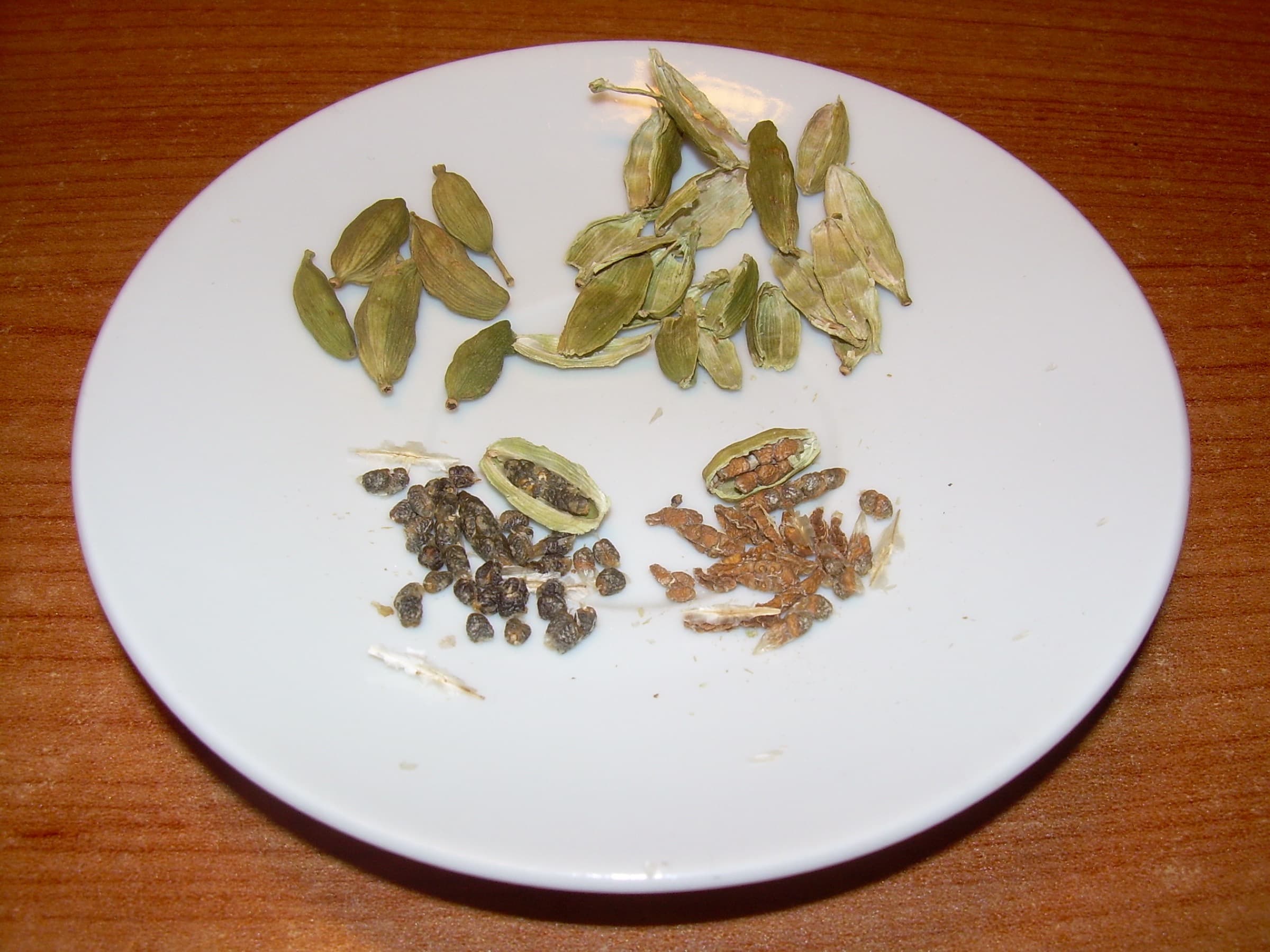 Green cardamom whole pods, empty husks, and loose dark and light seeds on a white plate