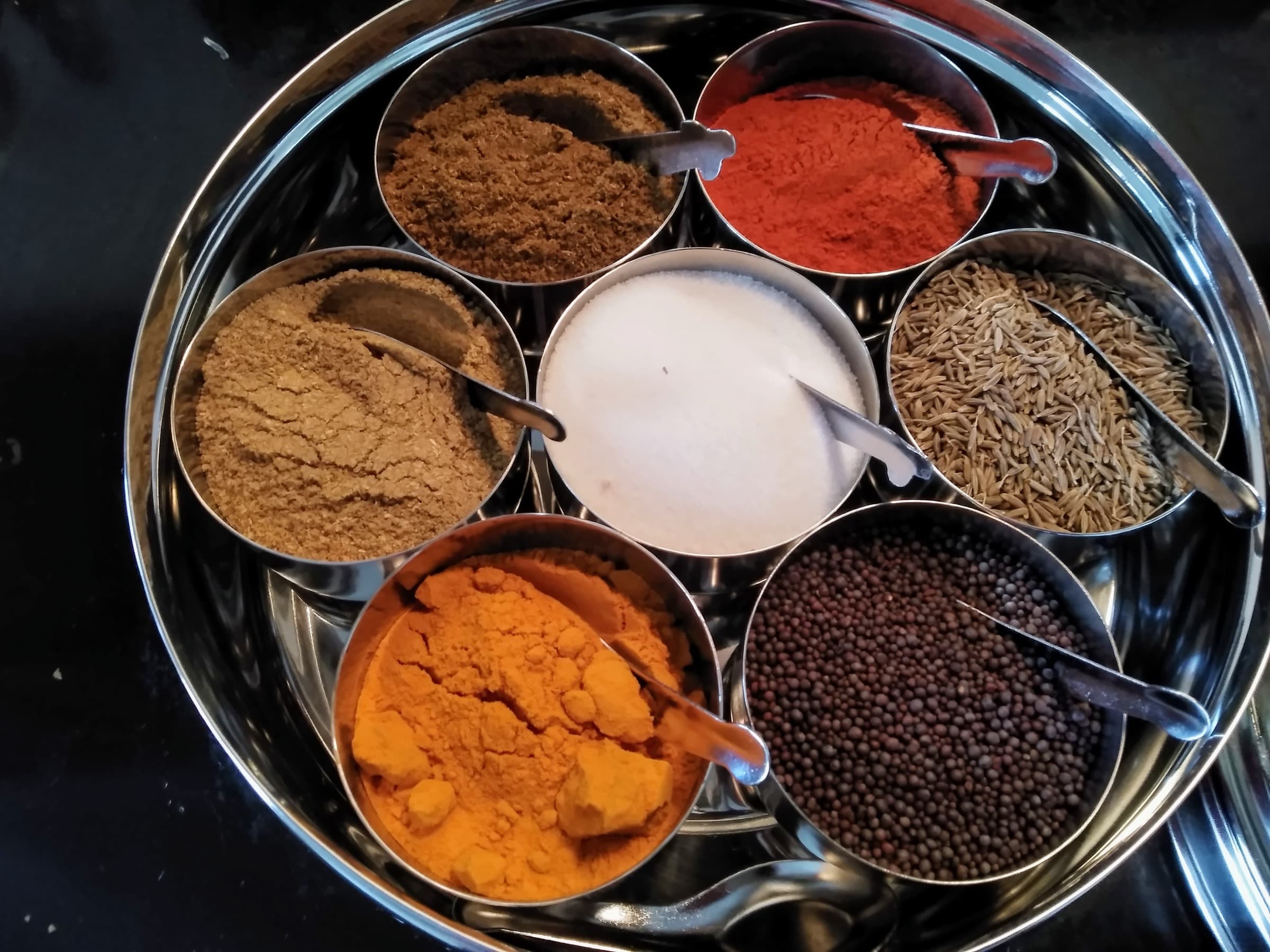 Open stainless masala dabba with seven spice cups and small spoons on a dark surface