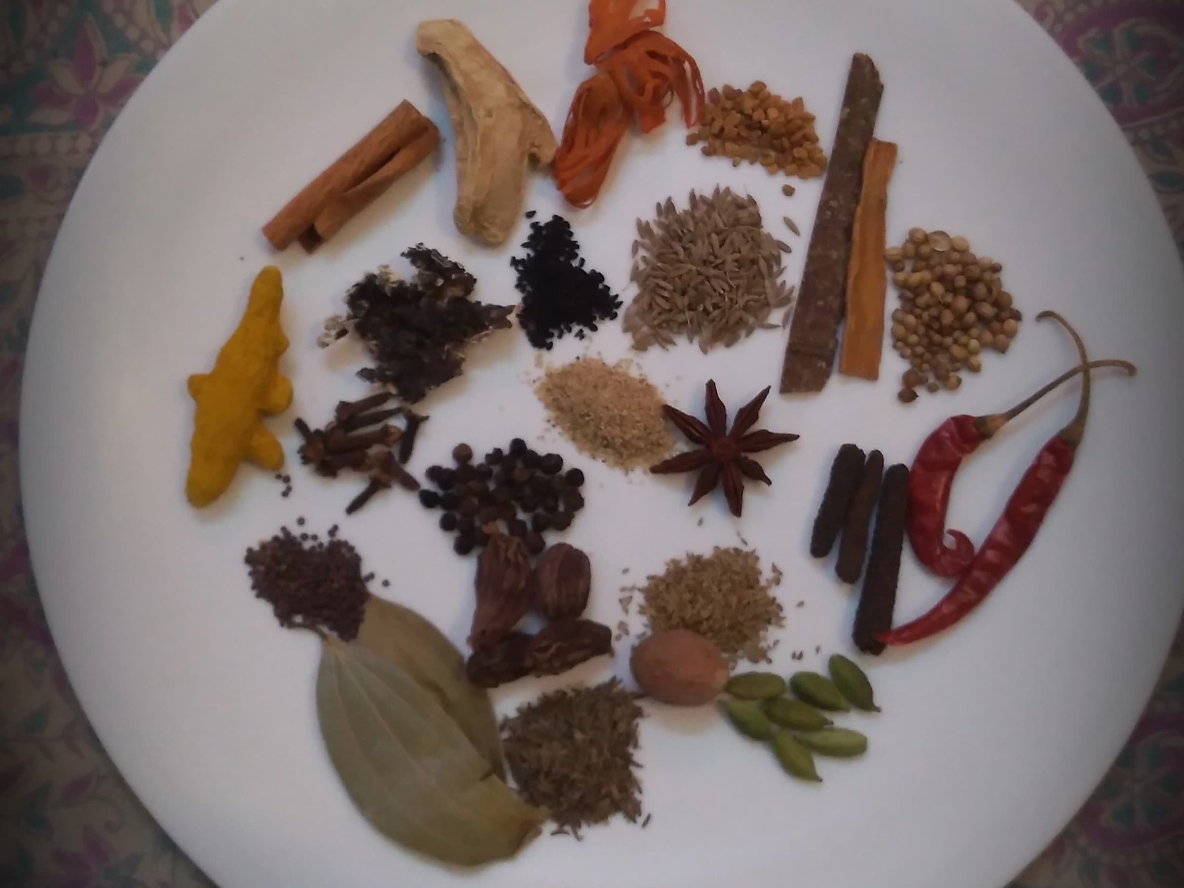 Assorted whole Indian spices on a white plate including cardamom, cinnamon, chilies, and cloves
